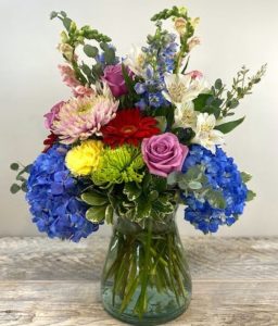A blooming assortment of blue hydranga, spider mums, lavender roses, alstroemeria, blue delphinium and pink snap dragon arranged in a glass conch vase